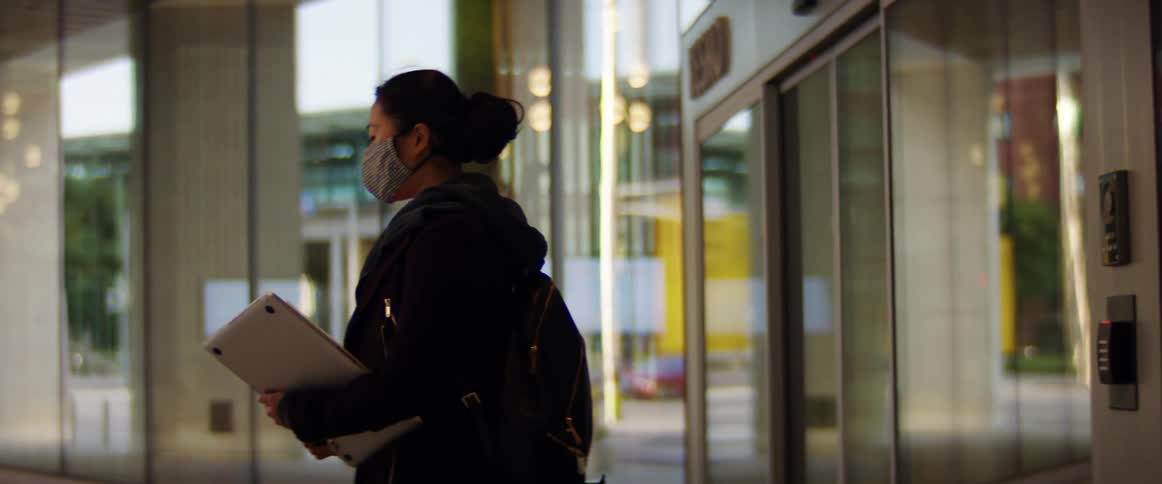 A woman wears a face mask indoors carrying a laptop