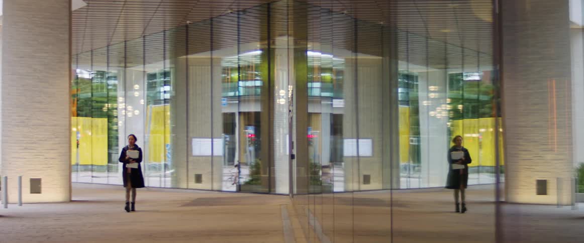 A woman with a trench coat walking next to a large glass window with the reflection of the city