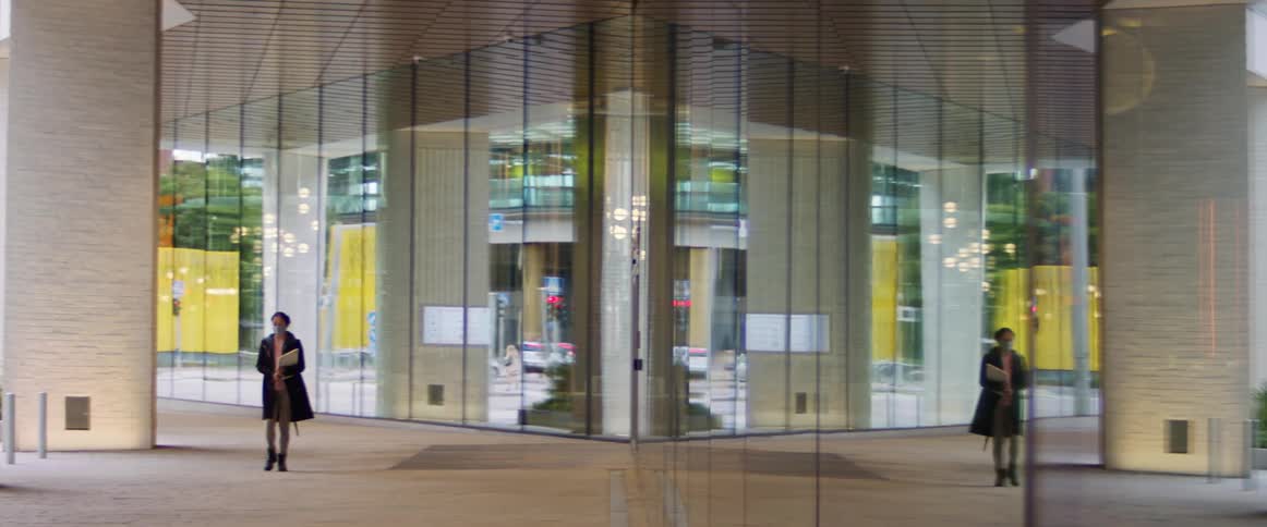 A woman in a medical mask walks in front of a large building while carrying a laptop
