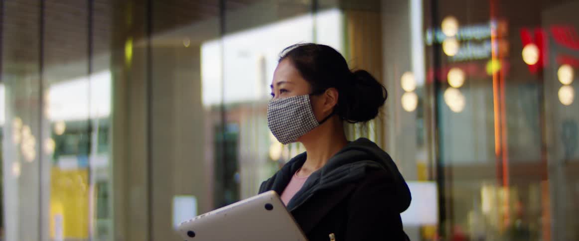 A woman walking down a city street carrying a laptop and wearing a mask