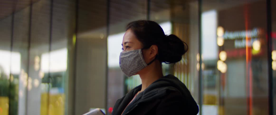 Woman wearing a covid mask holds a laptop while standing in front of some retail stores