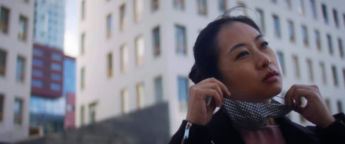 Woman wearing a mask a black jacket and pink top walks in front of some tall buildings and takes her mask off looking in the distance