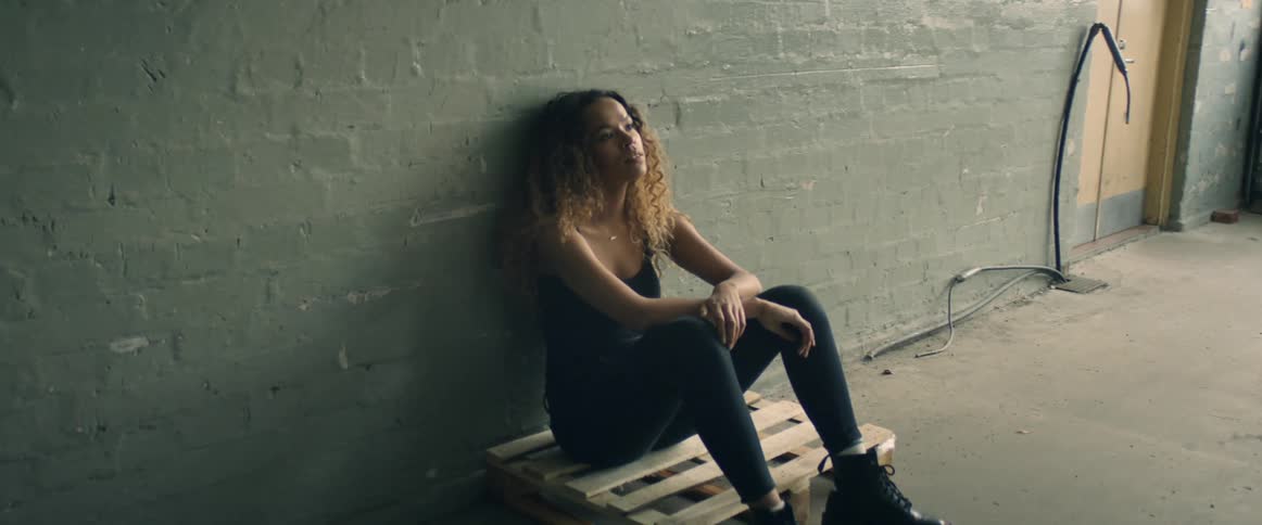 A woman looks bored while sitting on a small pallet in an empty room in a building