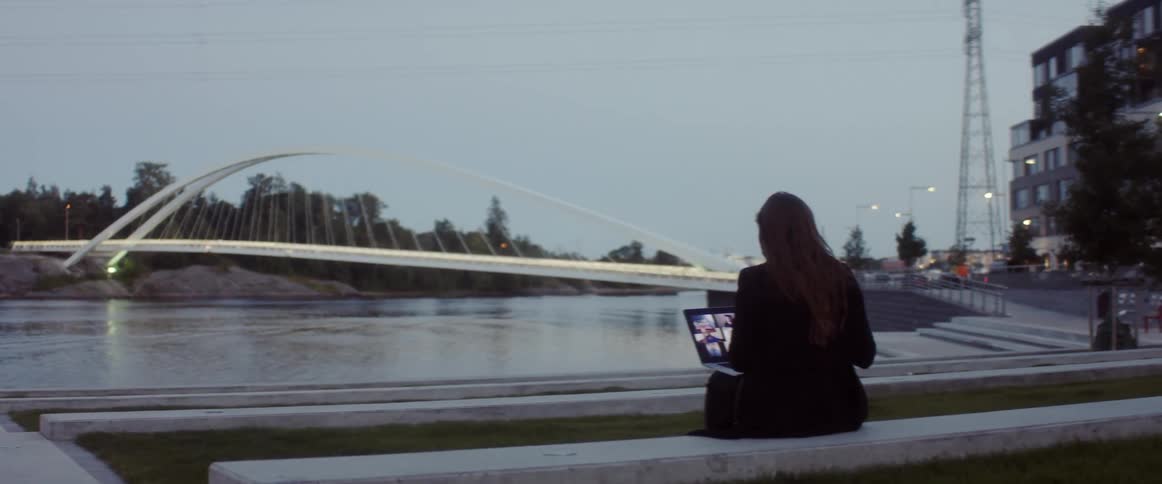 A woman sits on a bench with a laptop in front of water with a white bridge trees and buildings behind her