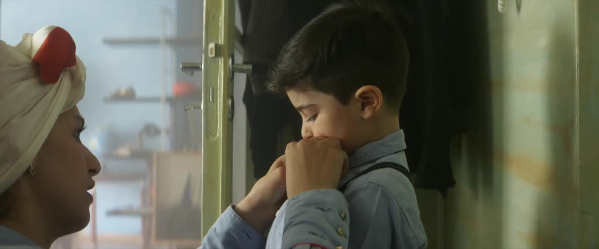 A woman kneels to tie the bow tie of her young son as he talks to her