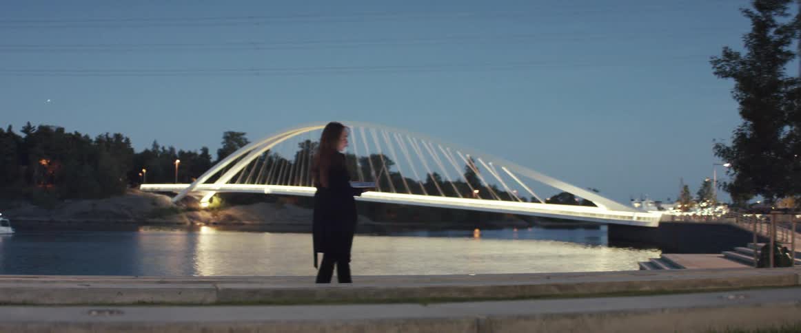 Woman wearing a black jacket and pants with a laptop and hair blowing in the wind places in front of a concrete pond with steps leading into it and a bridge lit up behind her and trees blowing in the wind