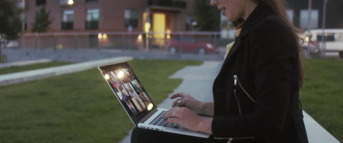 Woman sits on bench in fenced in grassy area wearing glasses a yellow shirt a black jacket pants and rings having a meeting with some people on her laptop with buildings cars and lights behind her