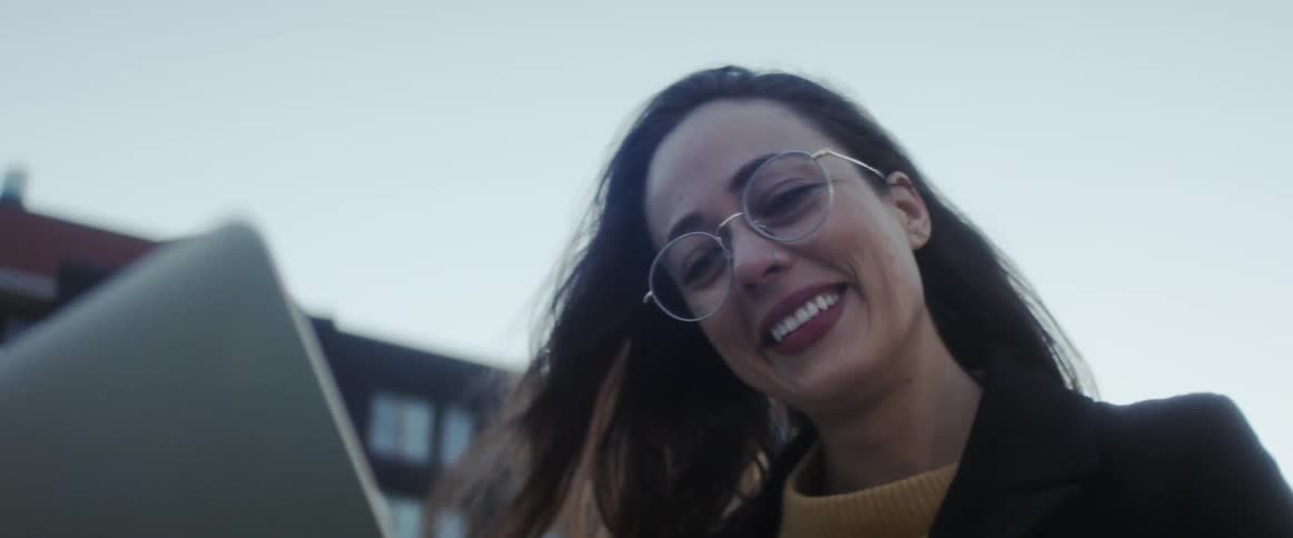 Woman wearing glasses smiles and laughs while having a video chat on a laptop outdoors