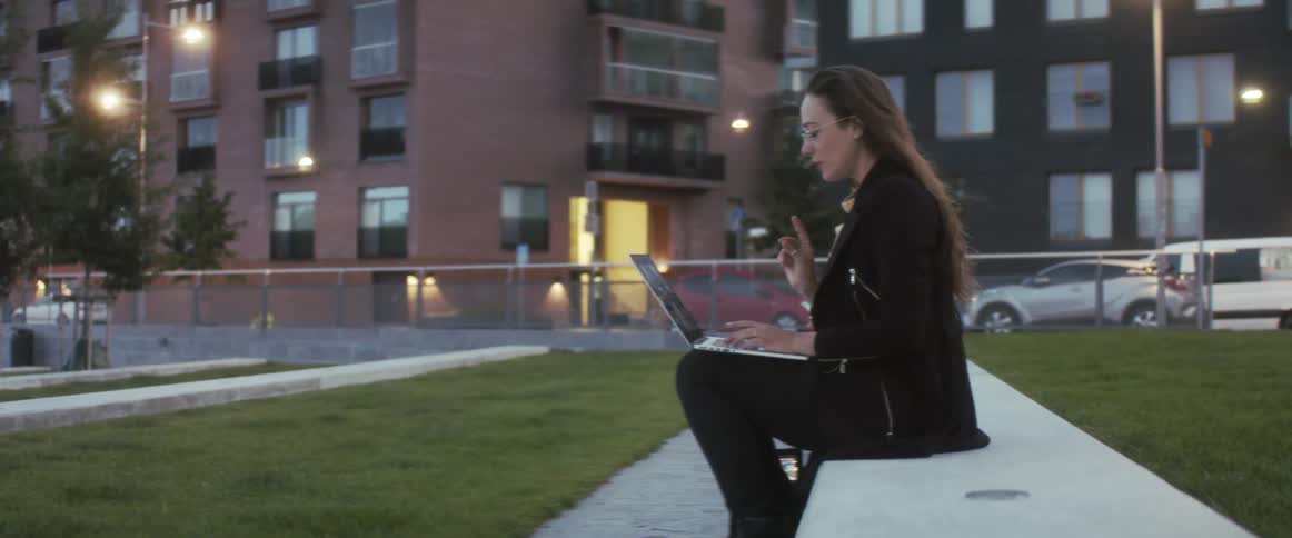 A woman sits on a ledge in a public park and talks to someone on her laptop