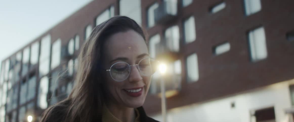 A woman with glasses standing and talking in a city with buildings the sky and clouds in the background