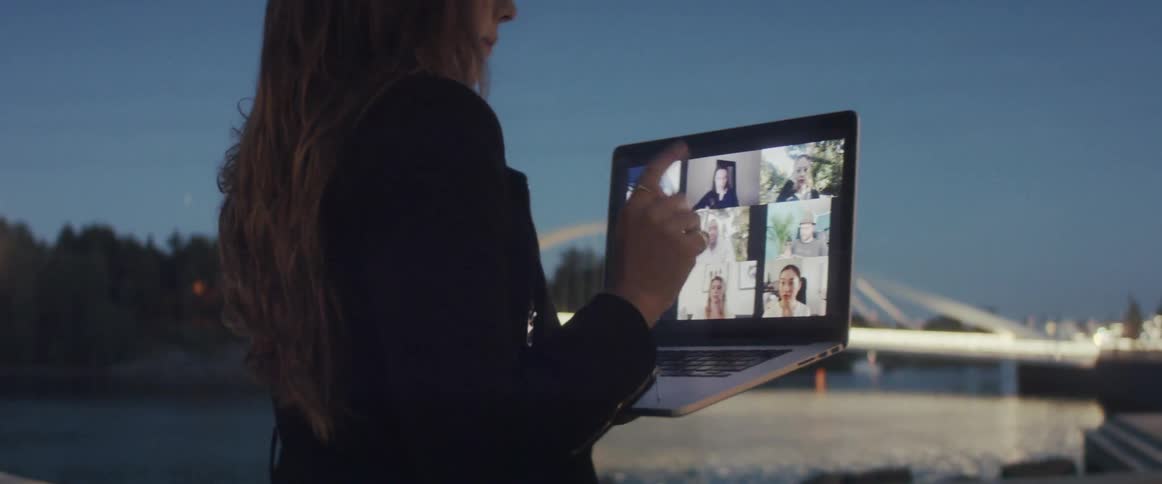 A woman in a suit standing on a riverbank participates in a video chat on a laptop she holds in front of her with one hand