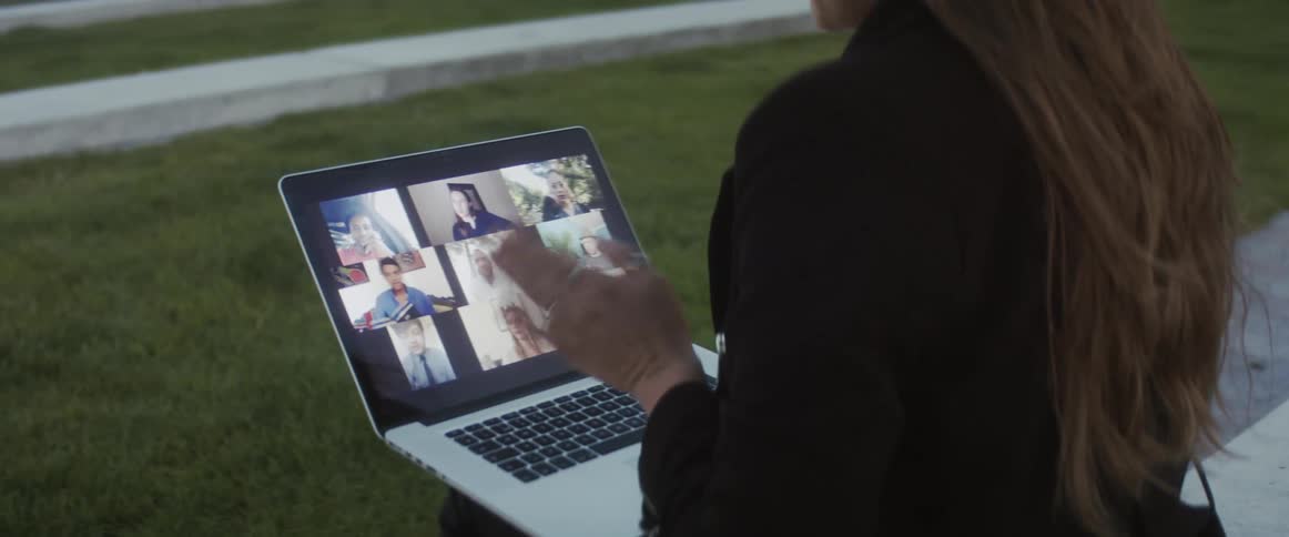 A woman sitting outdoors uses a laptop to participate in a video conference with several other people
