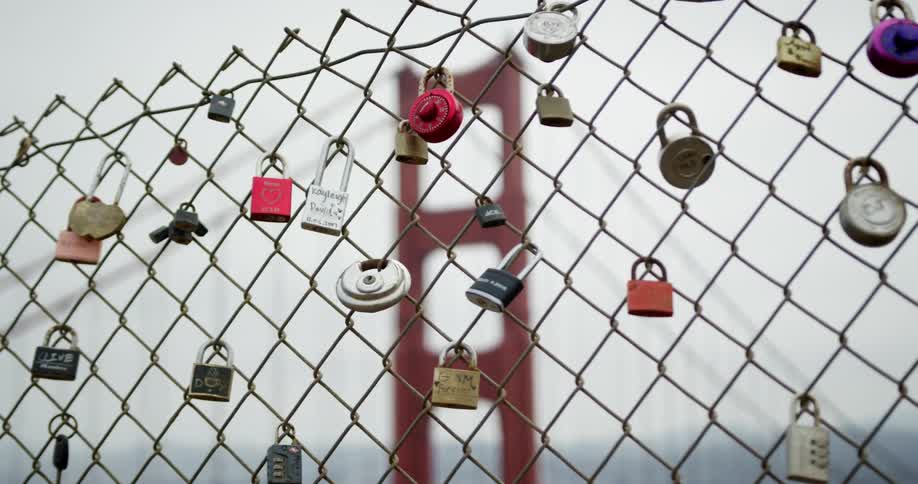 A fence with locks on it with a bridge the sky and clouds in the background