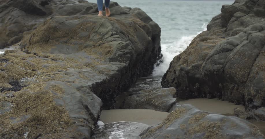 A woman walking across large rocks on the shore as the waves hit them