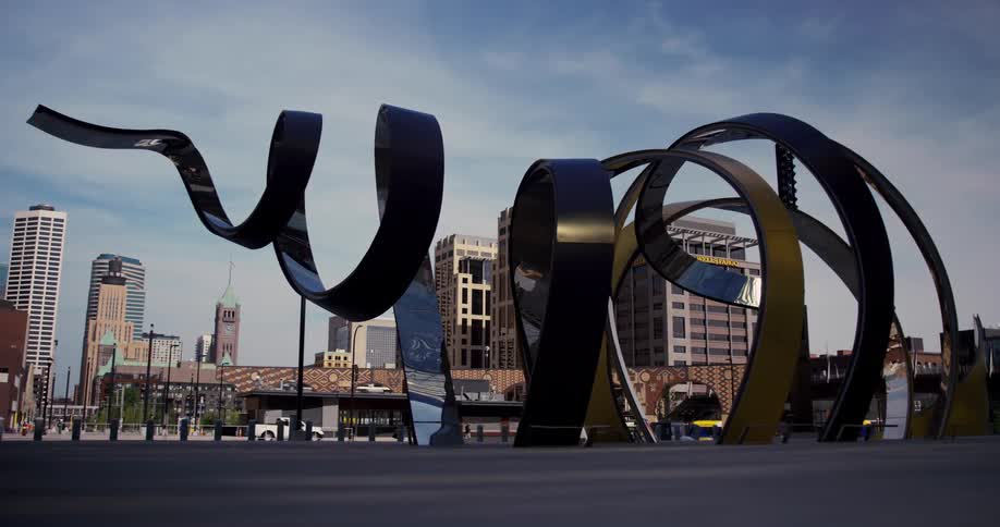Modern art statue in front of city street with truck passing on a sunny day with tall buildings in the background