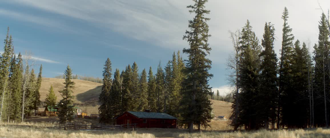 A cabin with a fence in the foreground with trees a hill the sky and clouds in the background