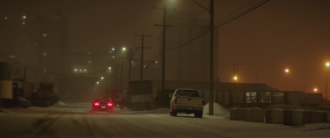 Car drives down city street lined with buildings power lines and street lights in the snow on a foggy night