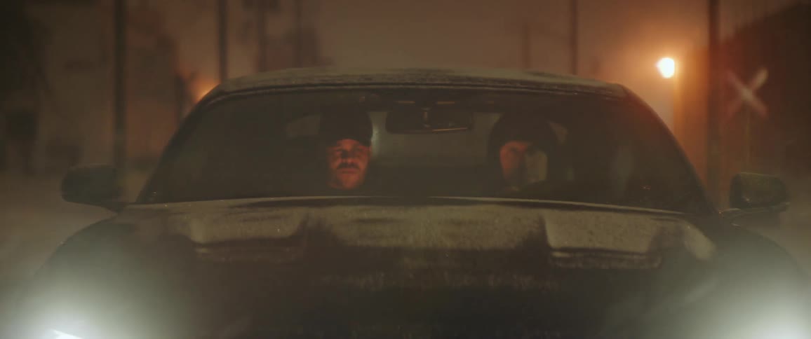 A pair of men speak while traveling through a city in a car on a snowy night