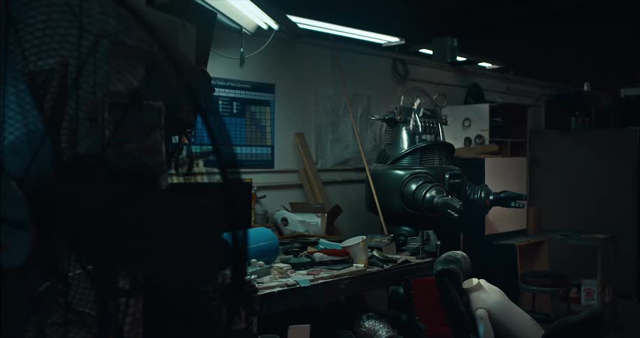 A lab with a flickering light contains a messy table with a large metal robot standing next to it and an incomplete robot on the floor with a flashing light on its arm