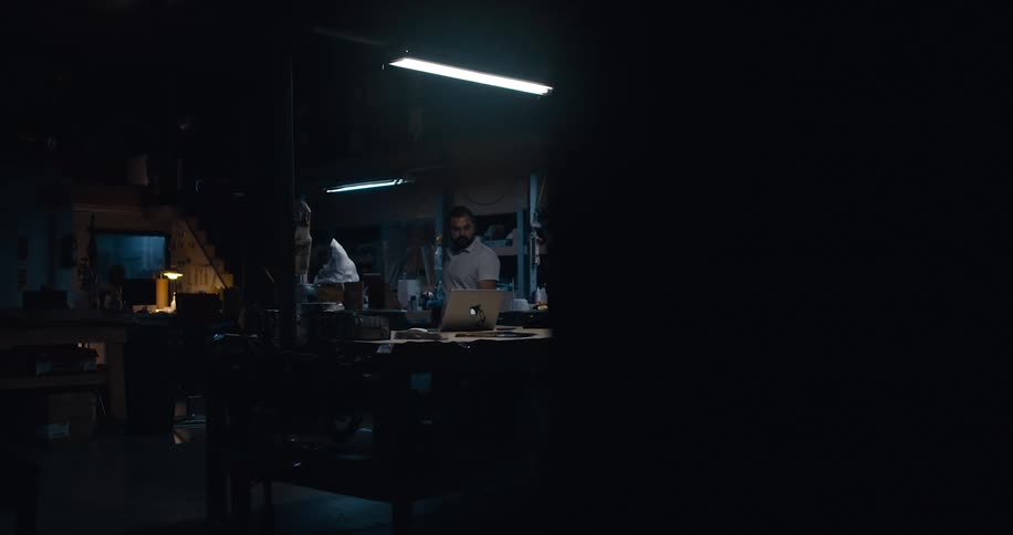 An male employee enters a dimly lit workroom and moves a mouse for the laptop sitting on a table