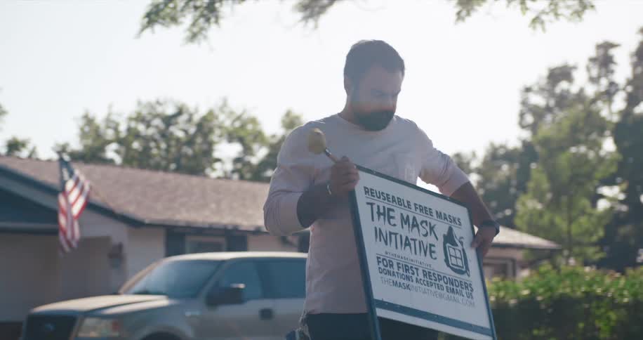 Man carries a sign about mask over to front lawn and places it there with hammer