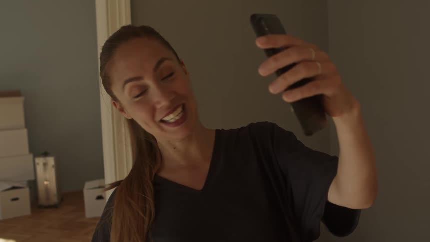A woman holds a phone as she speaks into it and shows the apartment she has just moved into with moving boxes visible in the background