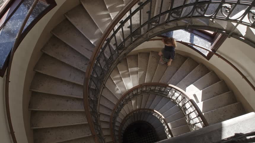 A female walks down a spiral staircase that has wood and iron railings with sunlight streaming through windows