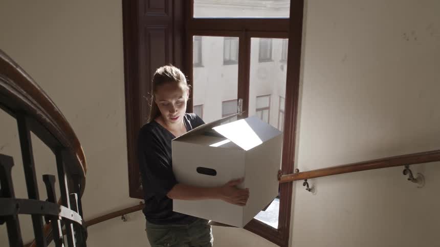 Woman carries white cardboard box up spiral staircase past window into hallway