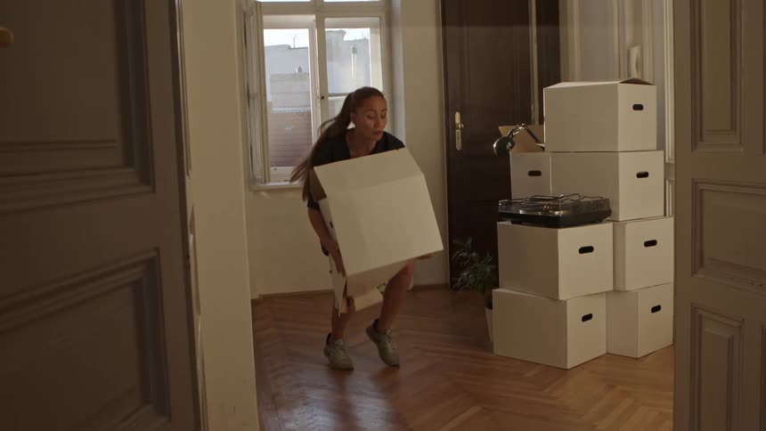 A woman carries a box into an apartment with the bottom of the box giving out as she gets frustrated