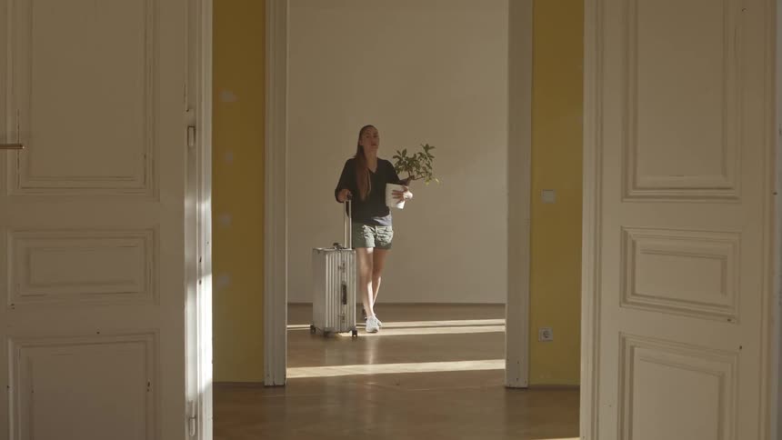 A woman walks into an empty home with a suitcase and potted plant