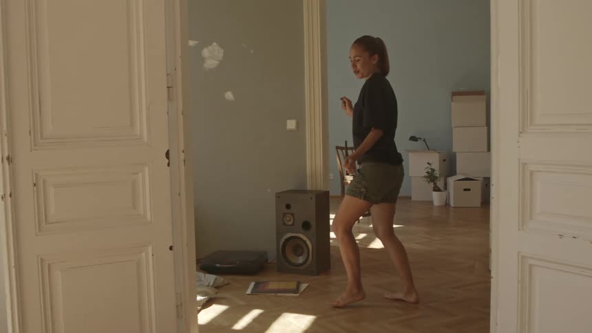 Woman dancing indoors in a room with chipped paint near a record player and speaker surrounded by moving and packing boxes