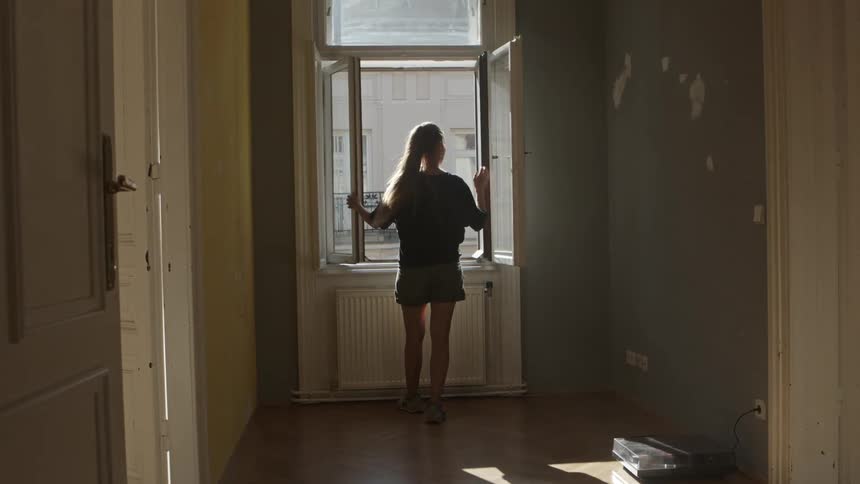 A woman stands and looks out a window at home and then closes the window up