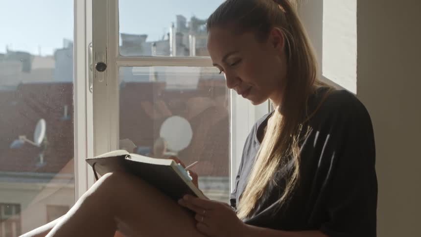 A young woman sits on a window balcony and writes in a book