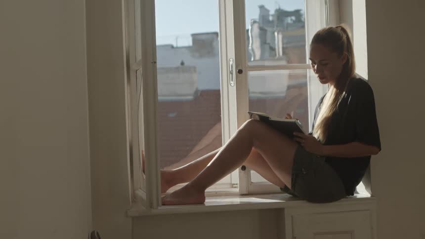 Woman with a ponytail a black t shirt and shorts sits on windowsill and writes in a notebook