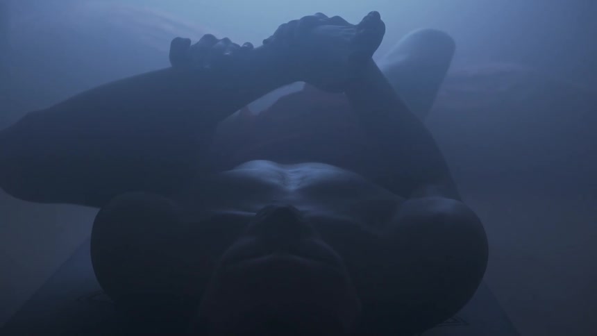 A shirtless man lies on a yoga mat while performing a yoga pose in a dimly lit room