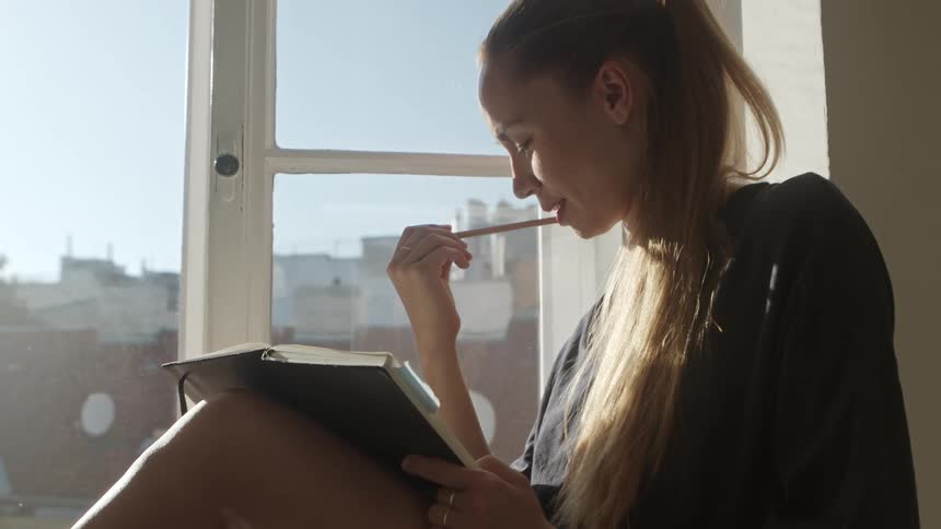 A woman is sitting by a window and reading a book