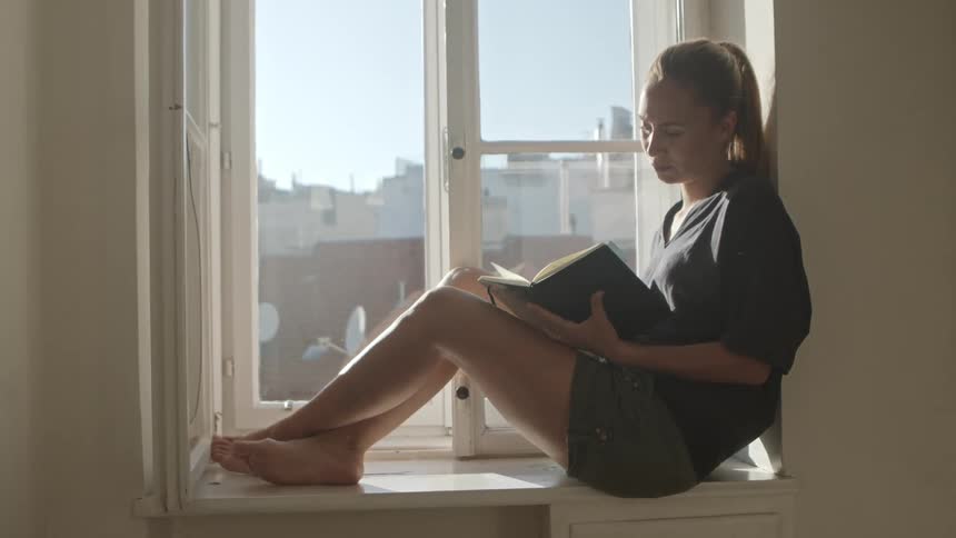 A young woman sits in a window at home and opens a book up