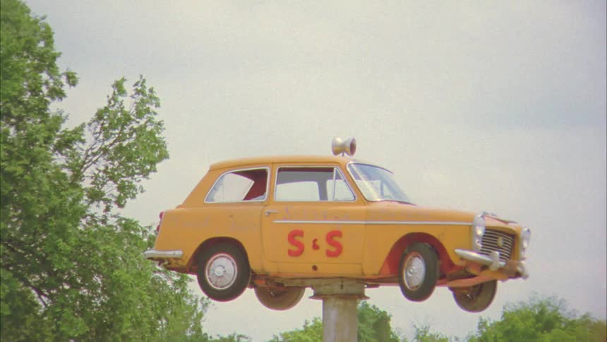 A compact orange car sits on top of a pole as it circles in front of trees in a town setting during the day
