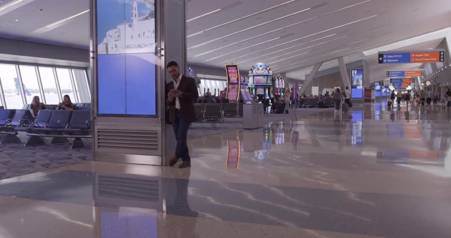 People walking and working in an airport and a man leans against a sign and looks at his cell phone