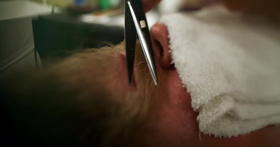A man lays in a barber chair having his beard trimmed with sharp scissors