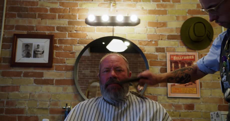 A seated man is having his hair and beard combed in a barbershop