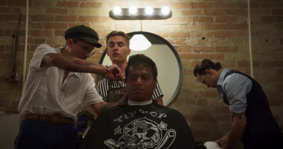 A man is demonstrating barbering techniques to two men with a seated customer inside a barbershop