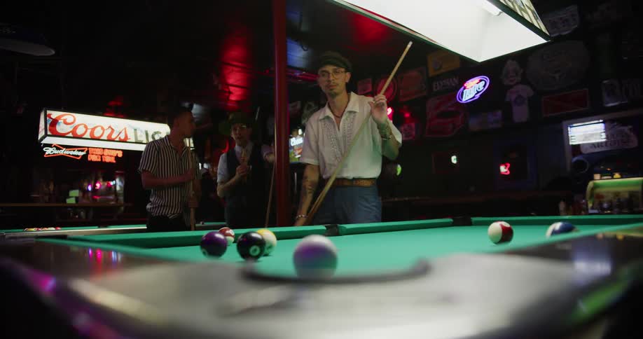 Inside of a bar a man takes aim and hits a billiard ball into a pocket on the table
