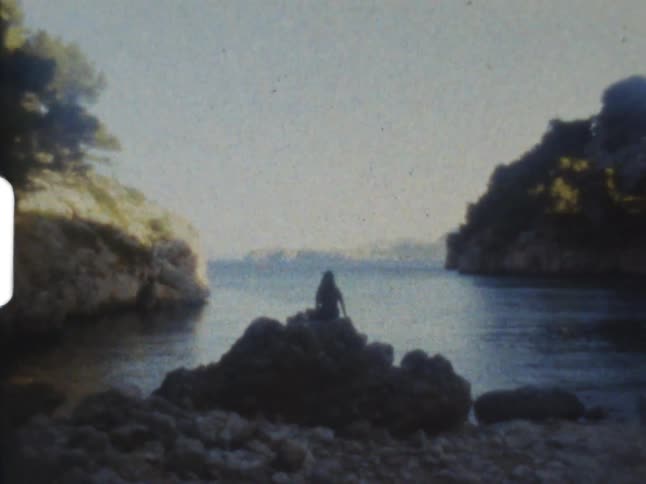Woman sits in shadow on a big rock on rocky beach looking out onto water with shore line in the distance and cliffs with tress to the side on 8mm