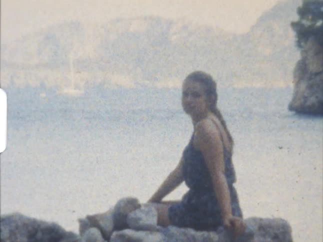 A young woman sitting on a rock formation on the coast looks back at the camera on 8mm