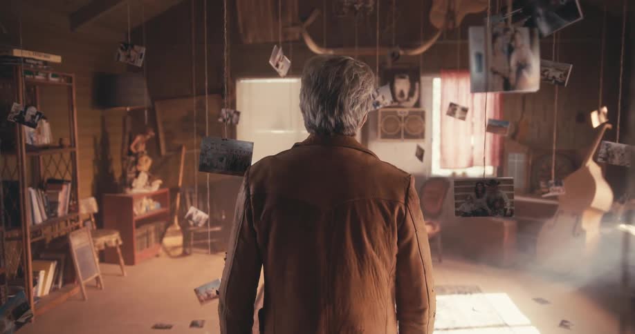 A man walks through an abandoned room with pictures hanging from the ceiling on strings and sun streaming through one window