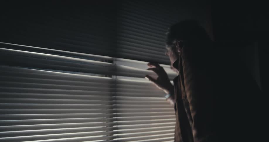 A man walks over his window and peeks through the blinds to see what is causing flashing lights outside