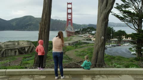 Mother and daughter stand while second daughter sits looking past park to ocean and bridge