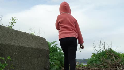 Girl in pink hoodie walks up incline until can view suspension bridge and opposite shore