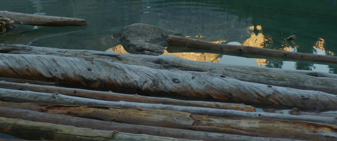 Ripples in water near collection of long tree logs fade revealing reflected mountains under clear blue sky